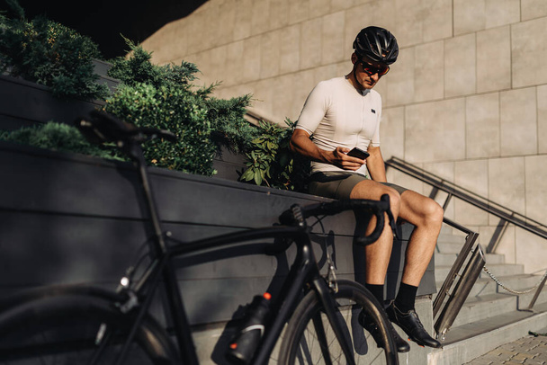 スポーツバイクの近くに屋外に座っている間、現代のスマートフォンを使用して安全ヘルメットや眼鏡で白人サイクリスト。生活のための技術. - 写真・画像