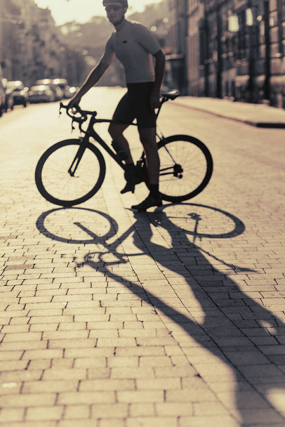Actieve blanke man in sportkleding, helm en bril rijden fiets op straat. Morgen zonlicht op de achtergrond. Schaduw op weg. - Foto, afbeelding