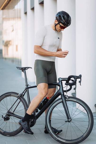Мускулистый спортсмен стоит на улице с велосипедом и держит в руках мультимедийный навигатор. Профессиональный велогонщик с использованием современного гаджета для обучения в городской местности. - Фото, изображение
