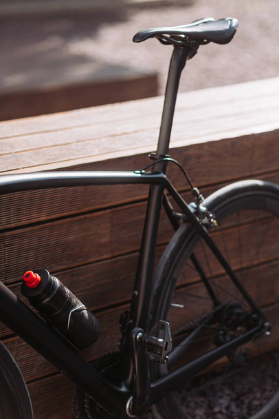 Close-up van zwarte sport fiets staan in de buurt van houten bank buiten. Professionele moderne uitrusting voor actieve training in frisse lucht. - Foto, afbeelding