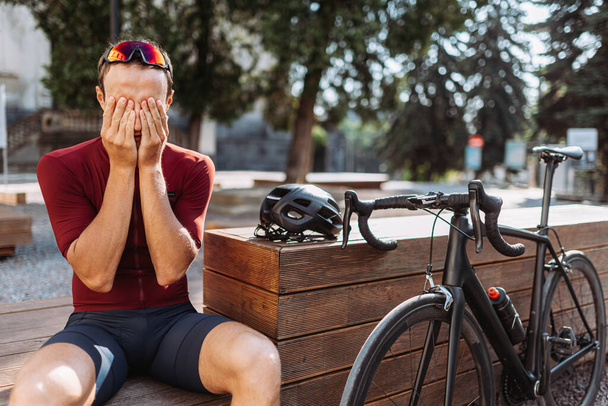 Уставший кавказский велогонщик сидит на деревянной скамейке и массирует лицо после долгой тренировки в шлеме и очках. Концепция выносливости, силы и отдыха. - Фото, изображение