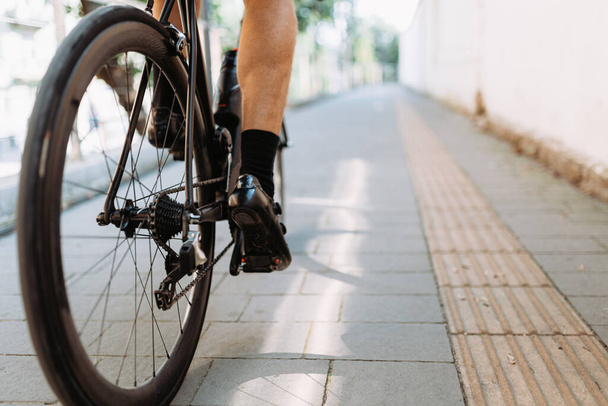 Gros plan du cycliste avec les jambes musclées chevauchant vélo noir sur la rue de la ville. Homme sportif en vêtements de sport cyclisme pendant la journée d'été en plein air. - Photo, image
