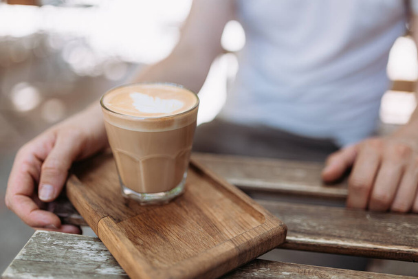Großaufnahme eines Mannes, der an einem Holztisch im Freien mit einer Tasse heißen, frisch gebrühten Kaffees sitzt. Männliche Person genießt Morgengetränk an frischer Luft. - Foto, Bild