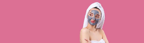 A woman with a gray clay cosmetic mask in a white towel on her head on a Pacific Pink background. - Фото, изображение