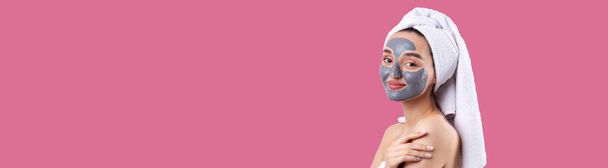 Een vrouw met een grijze klei cosmetische masker in een witte handdoek op haar hoofd op een Pacific Pink achtergrond. - Foto, afbeelding