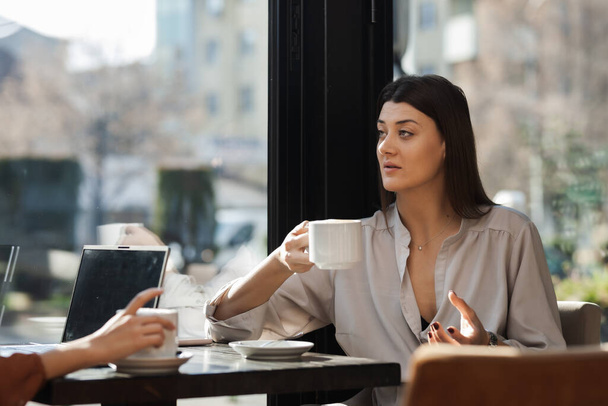 Two young business women in a cafe having one on one meeting. Friends after work talking gossiping and having coffee at a window table on a sunny day. - 写真・画像