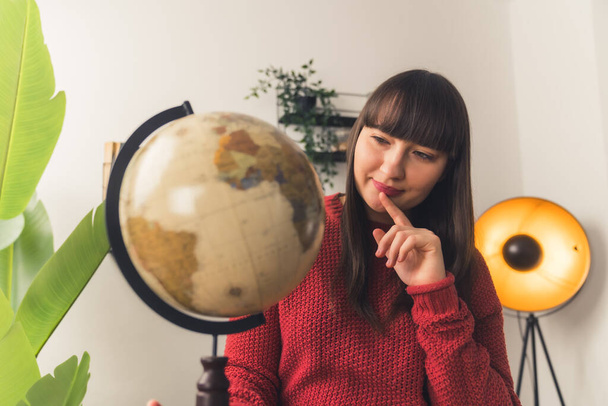 Casual caucasian woman in a red knitted sweater looking at an artificial globe and thinking about future travels. Indoor medium shot. - Photo, Image