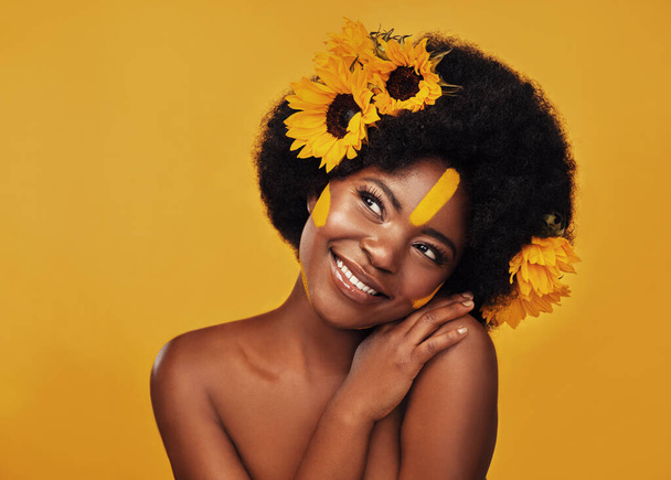 I wouldnt choose any other flower. Studio shot of a beautiful young woman smiling while posing with sunflowers in her hair against a mustard background. - Foto, Bild