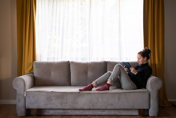 Молодая женщина использует цифровой планшет. Девушка что-то печатает, сидя на диване в современном и стильном интерьере - Фото, изображение