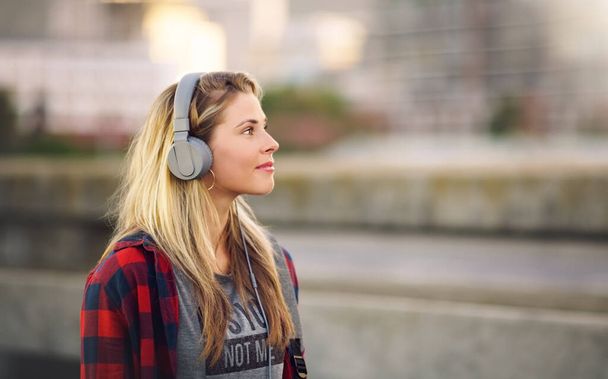 Ακούγοντας το ρυθμό. Τραβηγμένο πλάνο μιας ελκυστικής νεαρής γυναίκας που περπατάει στο δρόμο και ακούει μουσική μέσα από τα ακουστικά της.. - Φωτογραφία, εικόνα
