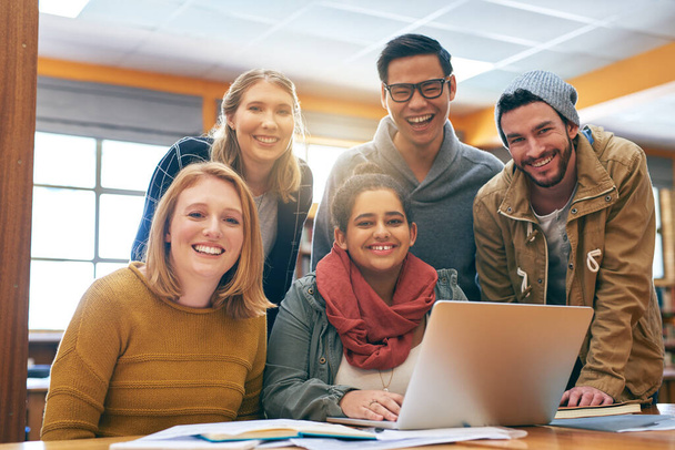 Het voelt een beetje beter als jullie samen studeren. Portret van een vrolijke jonge groep studenten die samenwerken met een laptop om te studeren voor examens in een bibliotheek. - Foto, afbeelding