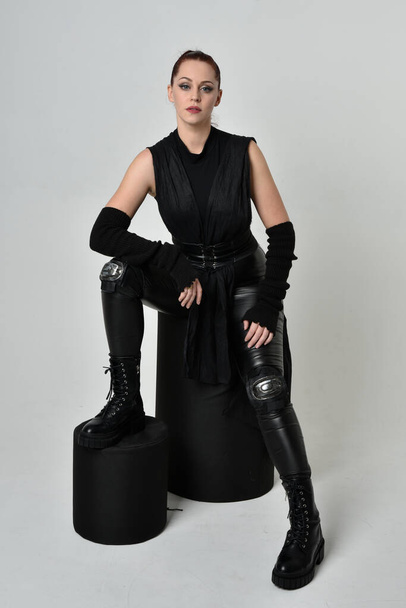 Full length portrait of pretty redhead female model wearing black futuristic scifi leather cloak costume. Dynamic sitting kneeling pose on white studio background. - Foto, afbeelding