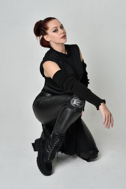 Volledig portret van mooie roodharige vrouwelijke model dragen zwarte futuristische scifi lederen mantel kostuum. Dynamisch zitten knielen pose op witte studio achtergrond. - Foto, afbeelding