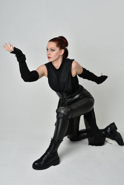 Full length portrait of pretty redhead female model wearing black futuristic scifi leather cloak costume. Dynamic sitting kneeling pose on white studio background. - Photo, Image