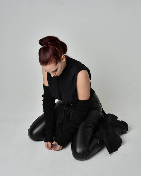 黒未来的なscifi革のマントの衣装を身に着けているかなり赤毛の女性モデルの完全な長さの肖像画。ダイナミック座位膝ポーズ上のホワイトスタジオの背景. - 写真・画像