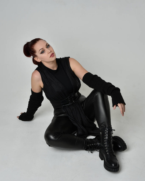 Teljes hosszúságú portré csinos vörös hajú női modellről fekete futurisztikus scifi bőr köpeny jelmezben. Dinamikus ülő térdelő póz fehér stúdió háttér. - Fotó, kép