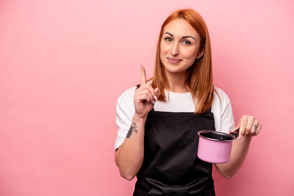 Joven mujer de cocina caucásica sosteniendo traductor aislado sobre fondo rosa mostrando el número uno con el dedo. - Foto, Imagen