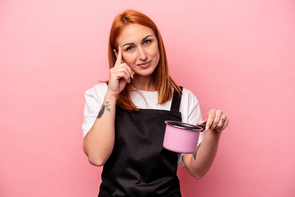 Joven mujer de cocina caucásica sosteniendo traductor aislado sobre fondo rosa apuntando templo con el dedo, pensando, centrado en una tarea. - Foto, Imagen