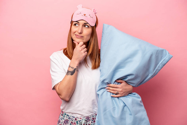 Young caucasian woman wearing a pajama holding pillow isolated on pink background looking sideways with doubtful and skeptical expression. - Valokuva, kuva