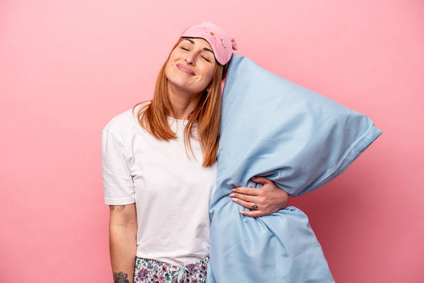 Young caucasian woman wearing a pajama holding pillow isolated on pink background dreaming of achieving goals and purposes - 写真・画像