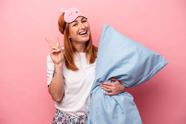Jeune femme caucasienne portant un pyjama tenant un oreiller isolé sur fond rose joyeux et insouciant montrant un symbole de paix avec les doigts. - Photo, image
