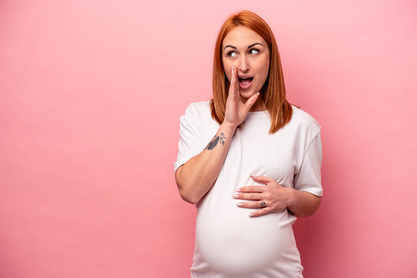 Joven mujer embarazada caucásica aislada sobre fondo rosa está diciendo una noticia secreta de frenado caliente y mirando a un lado - Foto, Imagen