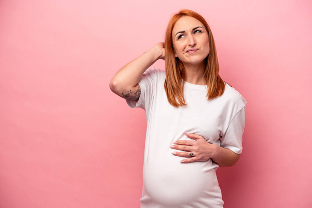 Junge kaukasische schwangere Frau isoliert auf rosa Hintergrund berührt Hinterkopf, denken und eine Wahl treffen. - Foto, Bild