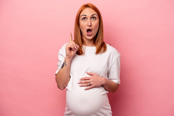 Junge kaukasische schwangere Frau isoliert auf rosa Hintergrund zeigt auf den Kopf mit geöffnetem Mund. - Foto, Bild