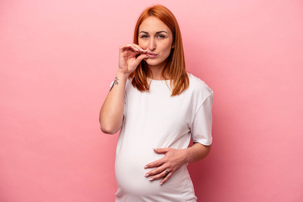 Young caucasian pregnant woman isolated on pink background with fingers on lips keeping a secret. - Photo, Image