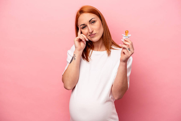 Young caucasian pregnant woman holding pacifier isolated on pink background pointing temple with finger, thinking, focused on a task. - Photo, Image