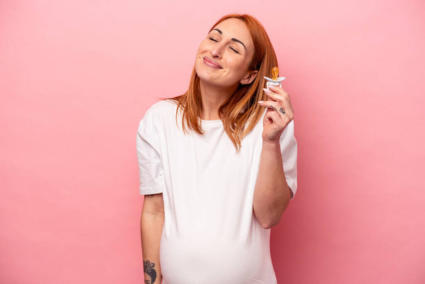 Young caucasian pregnant woman holding pacifier isolated on pink background dreaming of achieving goals and purposes - Foto, Imagen