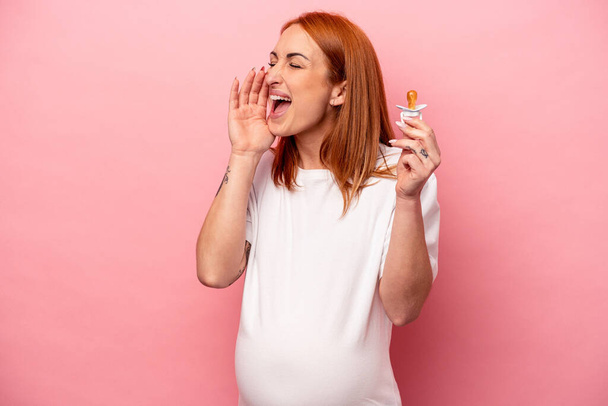 Young caucasian pregnant woman holding pacifier isolated on pink background shouting and holding palm near opened mouth. - Photo, image