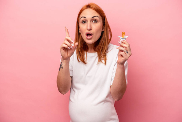 Young caucasian pregnant woman holding pacifier isolated on pink background having an idea, inspiration concept. - Photo, Image