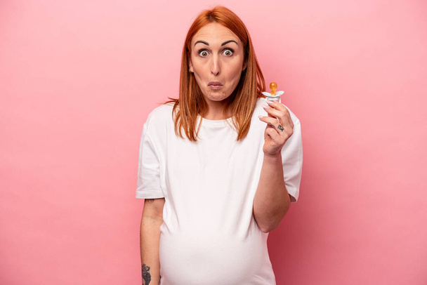 Young caucasian pregnant woman holding pacifier isolated on pink background shrugs shoulders and open eyes confused. - Fotoğraf, Görsel