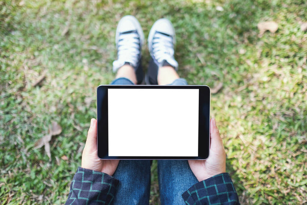 Top view mockup afbeelding van een vrouw houden en het gebruik van zwarte tablet pc met blanco witte desktop scherm tijdens het zitten in de buitenlucht - Foto, afbeelding