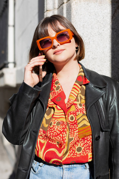 Portrait of young fashionable caucasian woman talking on the phone on the street. She is wearing a shirt with orange shades and matching sunglasses. Cool style and female fashion concept.  - Zdjęcie, obraz