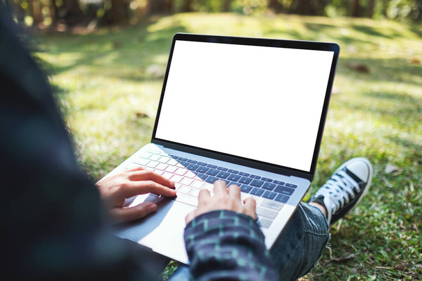 Mockup-Bild einer Frau, die auf Laptop mit leerem weißen Bildschirm tippt und im Freien mit Naturhintergrund sitzt - Foto, Bild