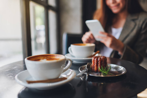 Крупним планом зображення азіатки, що тримає і використовує мобільний телефон з чашками кави і закусками на столі в кафе
 - Фото, зображення