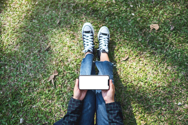 Top view mockup image of a woman holding black mobile phone with blank white screen while sitting in the outdoors - Valokuva, kuva