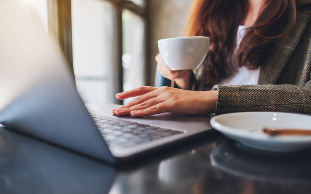 オフィスでコーヒーを飲みながらノートパソコンのタッチパッドで作業している女性のイメージを閉じる - 写真・画像