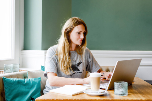 Egy fiatal nő portréja, aki okostelefon technológiával nézi a laptopot. Fiatal nő szabadúszó használ laptop számítógép dolgozik étteremben vagy hipster beltéri kávézó kávéval. Belföldi irodai koncepció. - Fotó, kép
