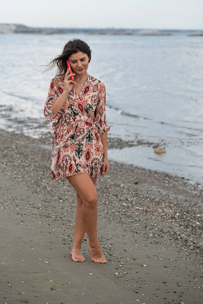 Mosolygó fiatal barna kaukázusi nő csevegett egy piros mobiltelefonnal a parton. Az emberek életmódjának fogalma - Fotó, kép