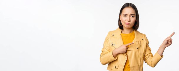 Déçu asiatique fille fronçant les sourcils, boudant bouleversé, pointant du doigt droite à la publicité, debout sur fond blanc - Photo, image