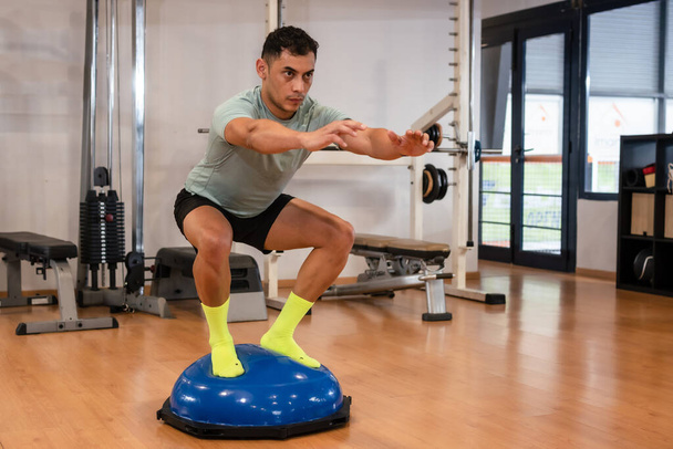 Портрет кавказца в спортзале, делающего балансовые упражнения для лодыжки на резиновой сфере, здорового и здорового образа жизни, занимающегося спортом - Фото, изображение