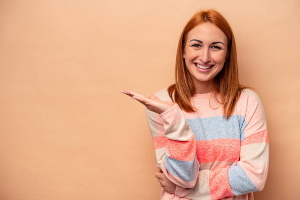 Young caucasian woman isolated on beige background showing a copy space on a palm and holding another hand on waist. - Photo, Image
