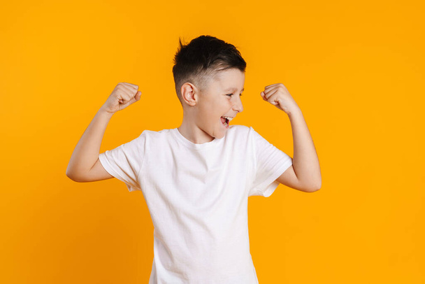 Brunette white boy in t-shirt smiling and making winner gesture isolated over yellow background - Fotó, kép