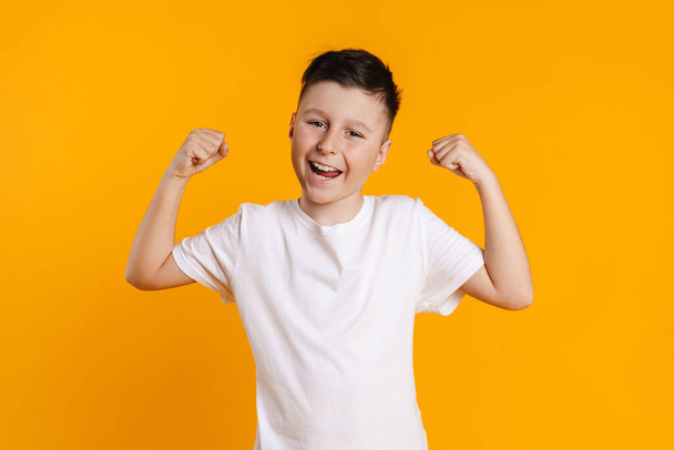 Brunette white boy in t-shirt smiling and making winner gesture isolated over yellow background - Zdjęcie, obraz