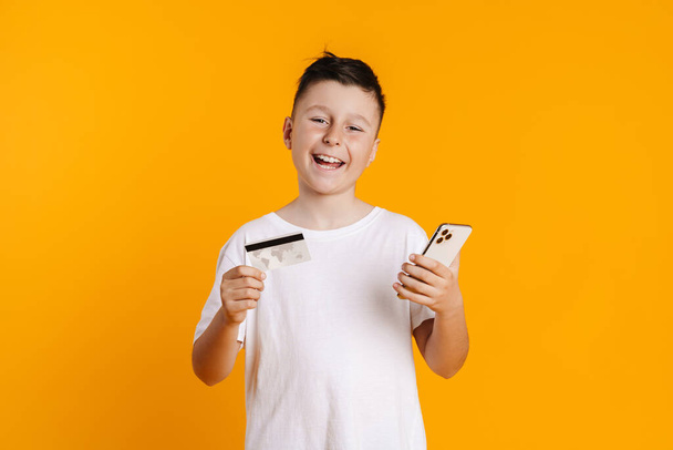 Brunette white boy posing with credit card and cellphone isolated over yellow background - Foto, Imagem