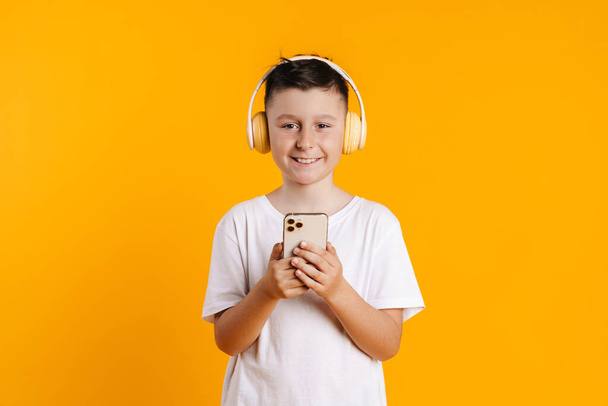 Brunette white boy smiling while using headphones and cellphone isolated over yellow background - Φωτογραφία, εικόνα