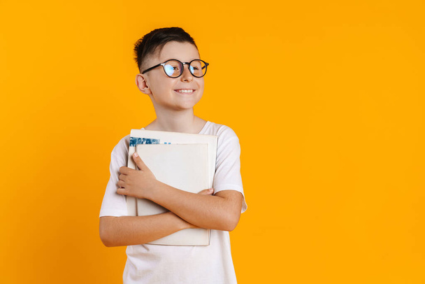 Brunette white boy in eyeglasses smiling while posing with exercise books isolated over yellow background - Foto, Imagen